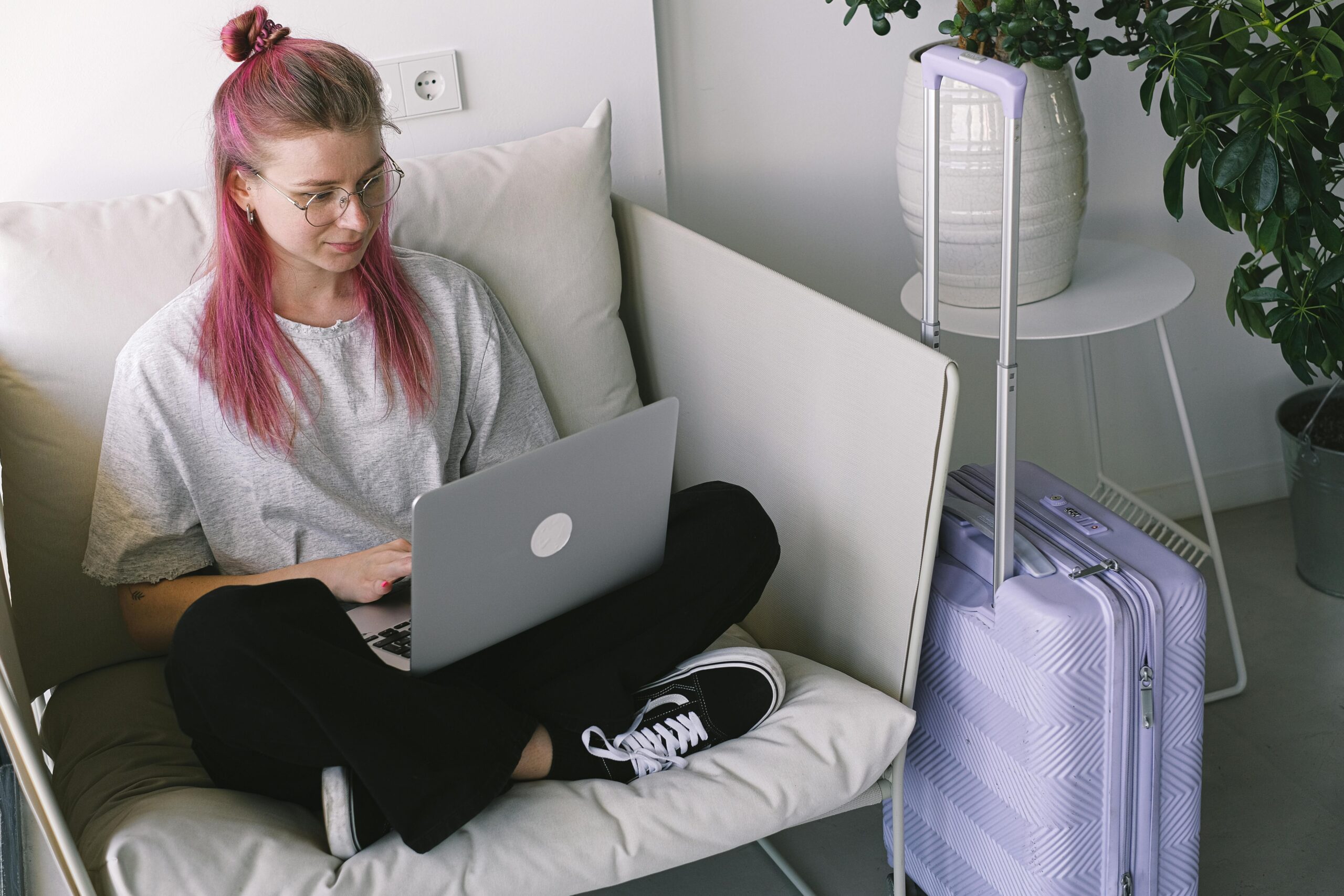 LÃ¤ssige junge Frau mit rosa Haaren arbeitet in gemÃ¼tlicher Innenumgebung am Laptop, Reisekoffer steht in der NÃ¤he.